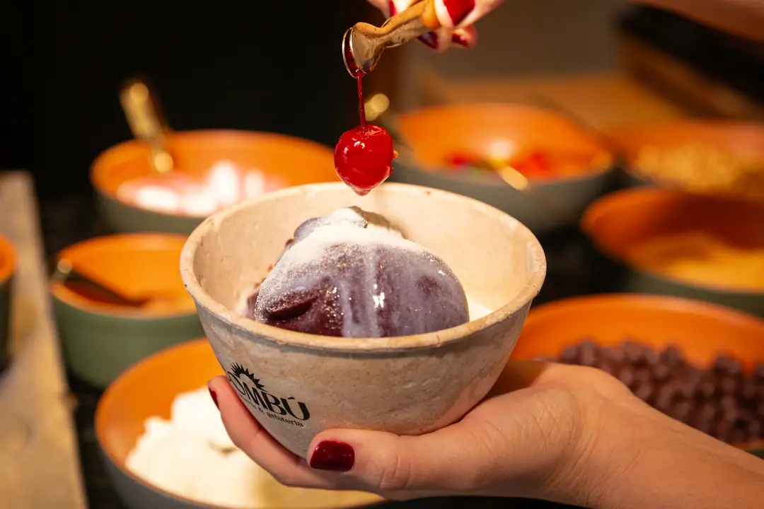 Cereja caindo sobre o açaí com cobertura, com os potes de cobertura ao fundo