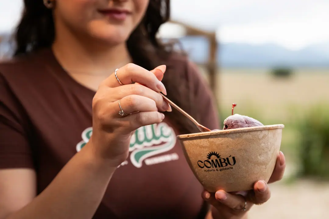 Mulher segurando o copo de açaí com cereja, com o campo e a serra ao fundo
