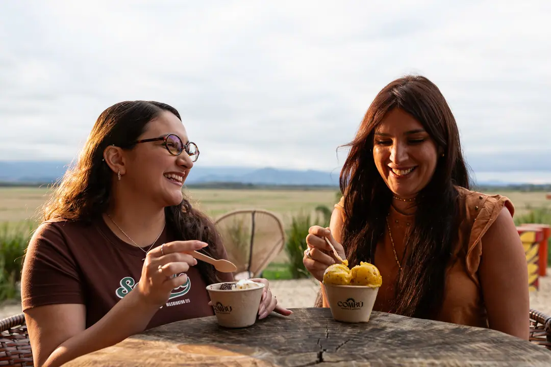 Duas amigas rindo numa mesa ao ar livre, com copos de sorvete e a Mantiqueira no horizonte