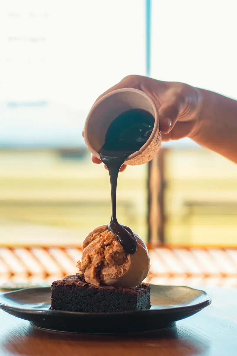Calda escura escorrendo sobre sorvete e brownie, com o deck ao fundo