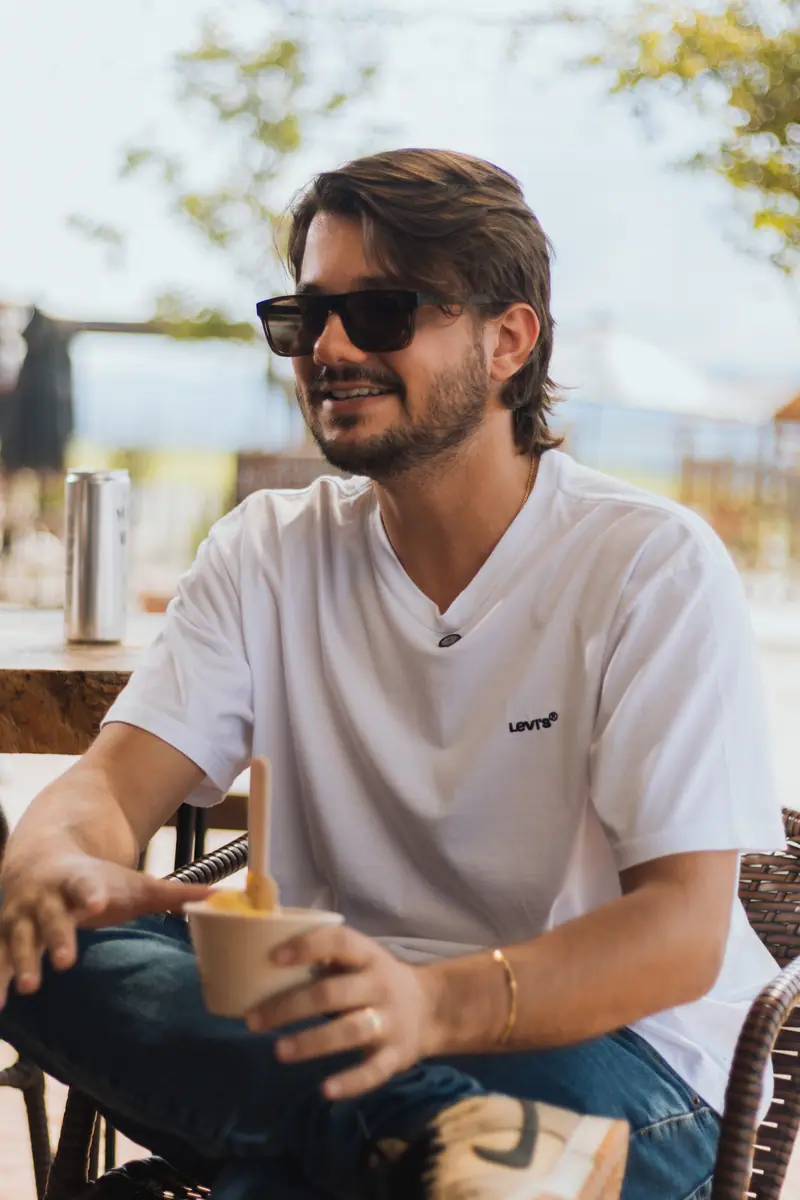 Rapaz sentado no deck, de óculos escuros, com um copo de sorvete na mão