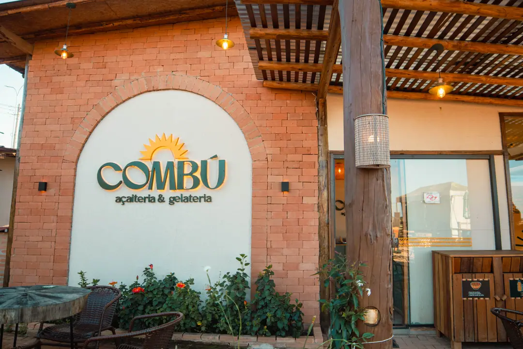 Fachada de tijolo com o arco branco e o logo da COMBÚ, jardim e pergolado