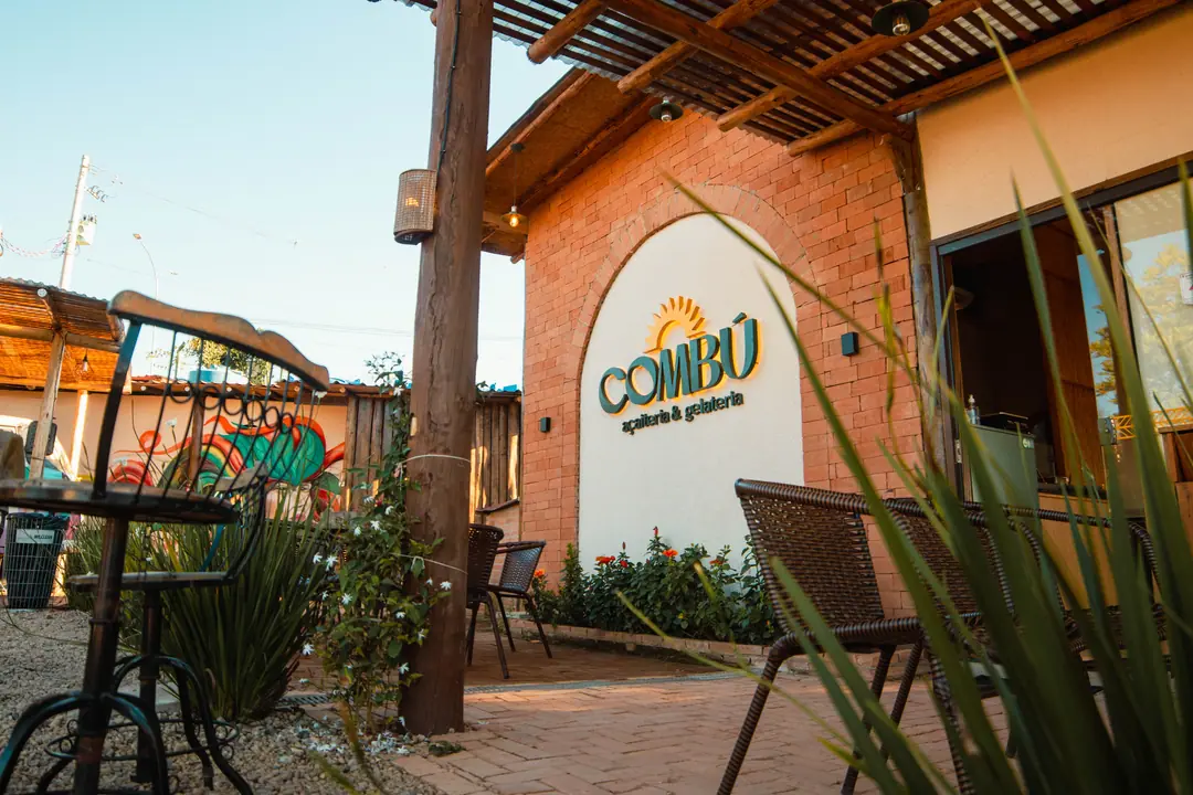 Entrada da COMBÚ vista do jardim, com o logo no arco e cadeiras de fibra ao sol