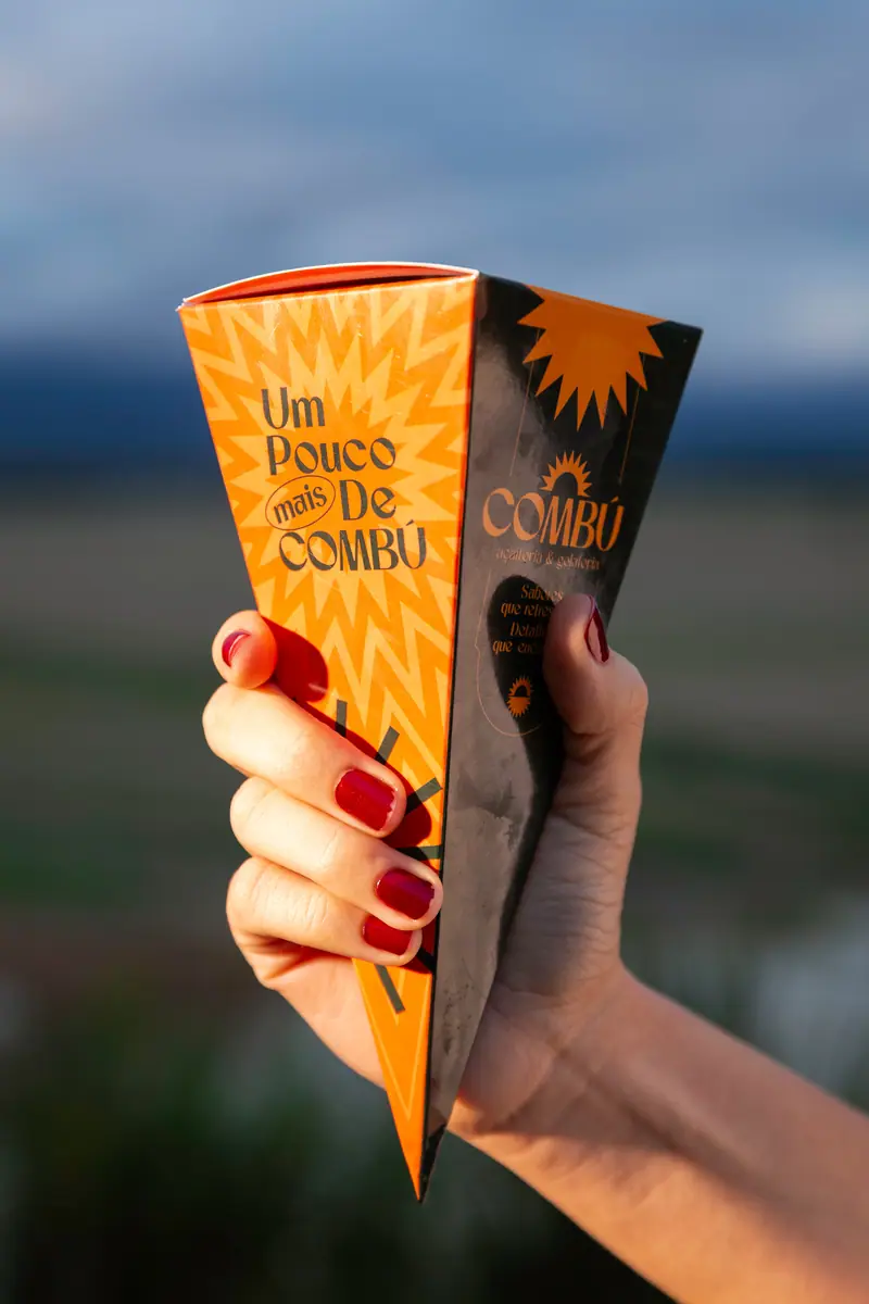Mão segurando a embalagem laranja da casquinha no fim de tarde, com a serra azulada ao fundo