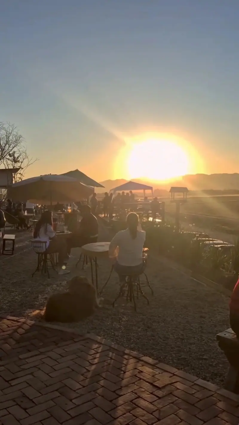 Sol se pondo atrás da serra, pessoas nas mesas ao ar livre e um cachorro deitado no cascalho